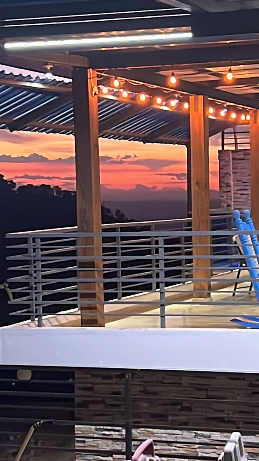A spacious terrace is highlighted by warm, outdoor lighting strung overhead. The railing offers a glimpse of the scenic landscape beyond, where vibrant colors of the sunset are visible in the sky, creating a peaceful atmosphere. A swimming pool is partially visible on the terrace.