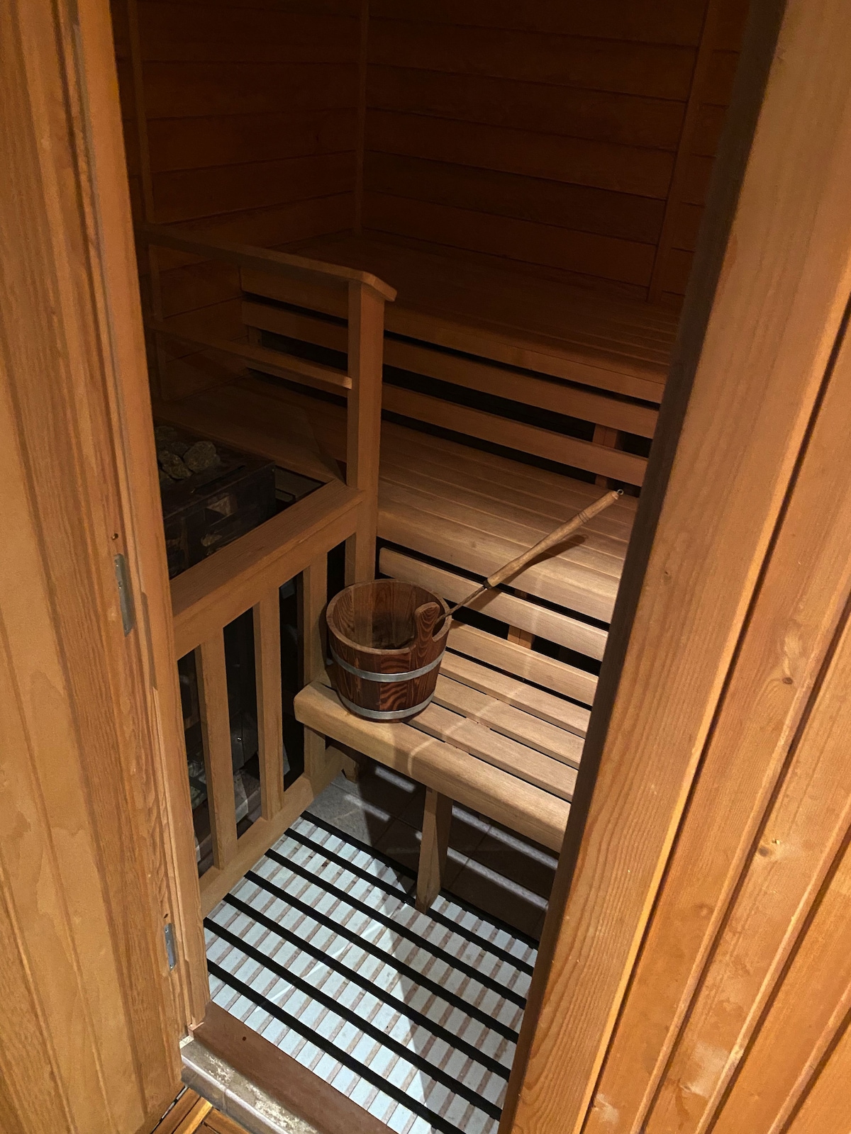 A wooden sauna interior is visible, featuring bench seating along the walls. A wooden bucket and a ladle are positioned on a nearby bench, indicating an area for water application. The floor is structured with slats, enhancing ventilation and moisture control.