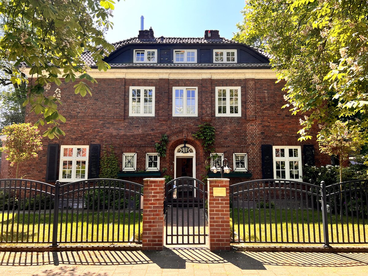 A stately villa is surrounded by lush greenery, featuring a brick façade and elegant windows. The entrance is marked by a wrought-iron gate, with a manicured garden visible in the foreground. Architectural details highlight the home’s classic charm.