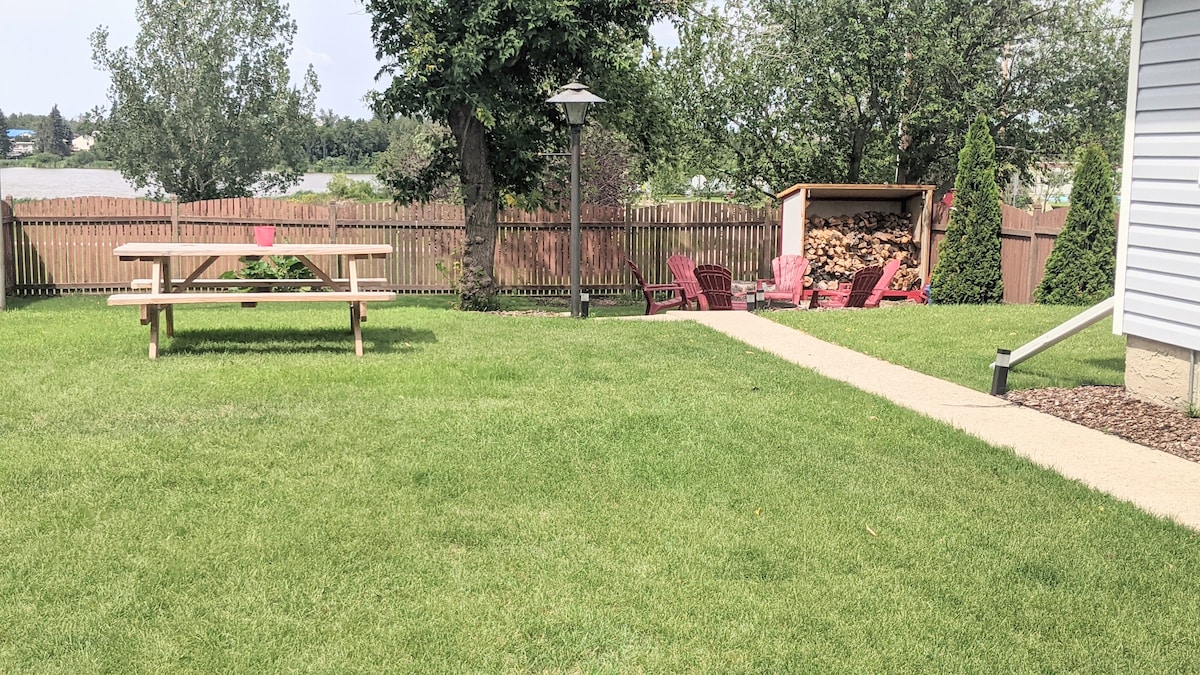 A spacious yard is visible, featuring a picnic table and a fire pit area with red chairs. A wood storage shed is placed against the fence, surrounded by lush green grass and trees. The serene backdrop includes a glimpse of the nearby lake.