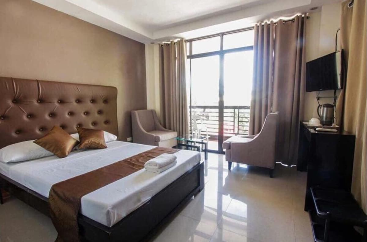 A well-appointed room featuring a spacious bed with a padded headboard and neatly arranged linens. Two comfortable armchairs are positioned by a window, allowing for natural light. A flat-screen TV is mounted on the wall, and a small table is seen beside the seating area.