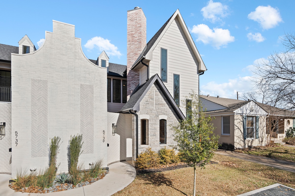 The exterior of the home features a blend of textures, with a light-colored brick facade and natural stone elements. A pathway lined with shrubs leads to the entrance, and a gracefully manicured lawn complements the structure's inviting appearance.