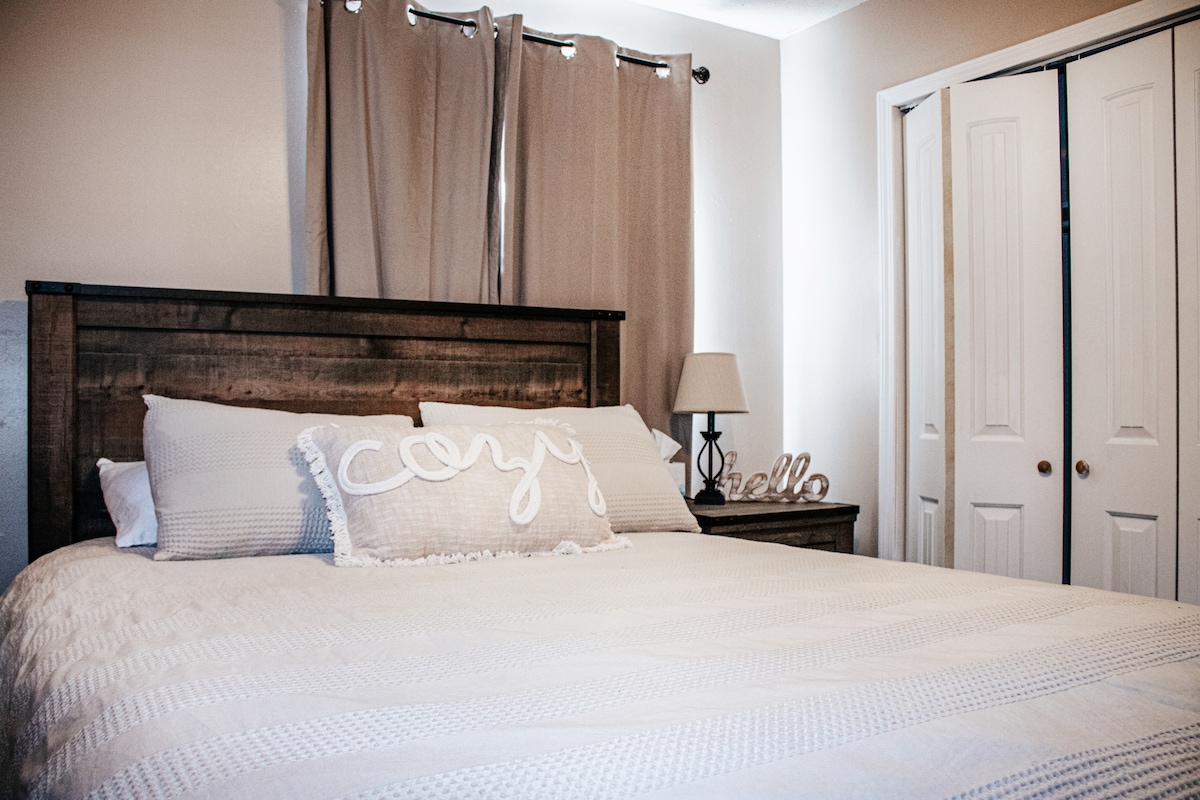 A spacious bed is centered in the room, adorned with a textured white blanket and a decorative 'Cozy' pillow. Soft natural light enters through window coverings, and a bedside lamp provides additional illumination. A nearby closet with double doors is visible.