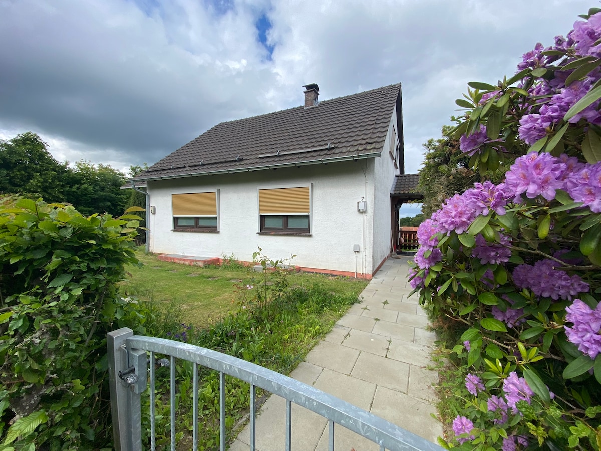 A charming house with a sloped roof and white exterior is framed by vibrant blooming rhododendrons. A paved pathway leads from a gate to the entrance, providing a welcoming approach surrounded by greenery.