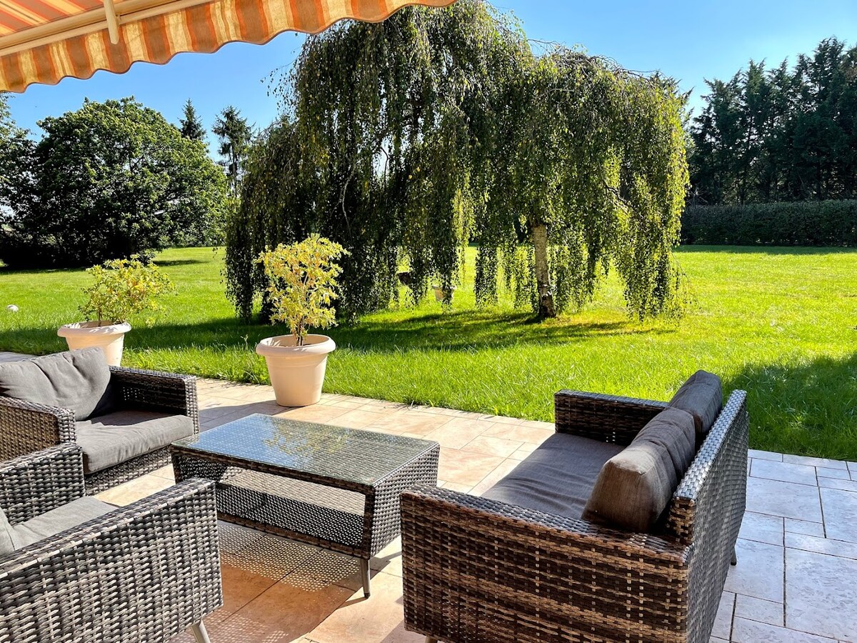 A shaded terrace area features comfortable seating with wicker sofas and a glass-top table. A sprawling green lawn stretches beyond, adorned with a weeping willow tree and surrounding foliage creating a serene outdoor environment.