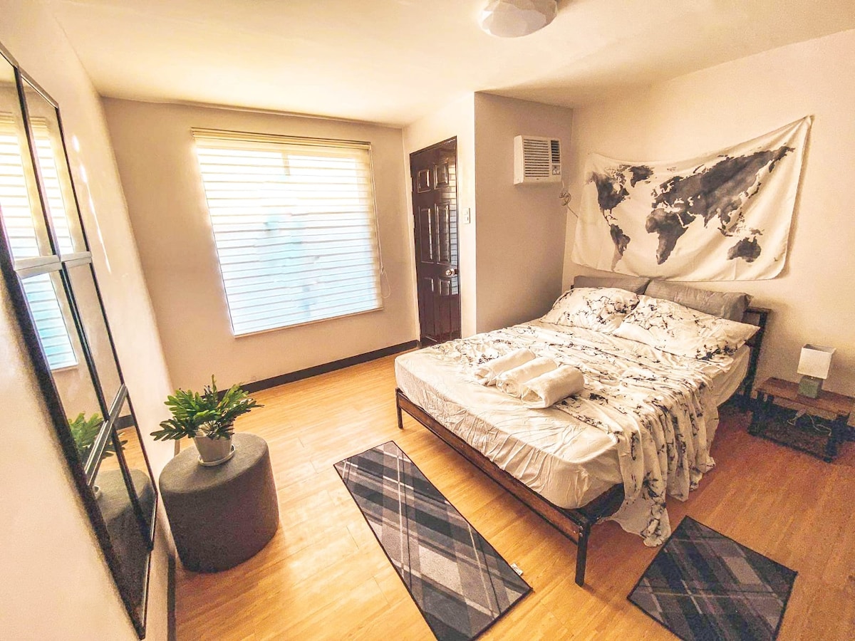 A well-furnished bedroom features a spacious bed adorned with a world map motif blanket. Natural light filters through a window with blinds, illuminating the warm wood floor. A minimalist mirror and a small round plant stand add to the room's serene ambiance, complemented by a neutral color palette.