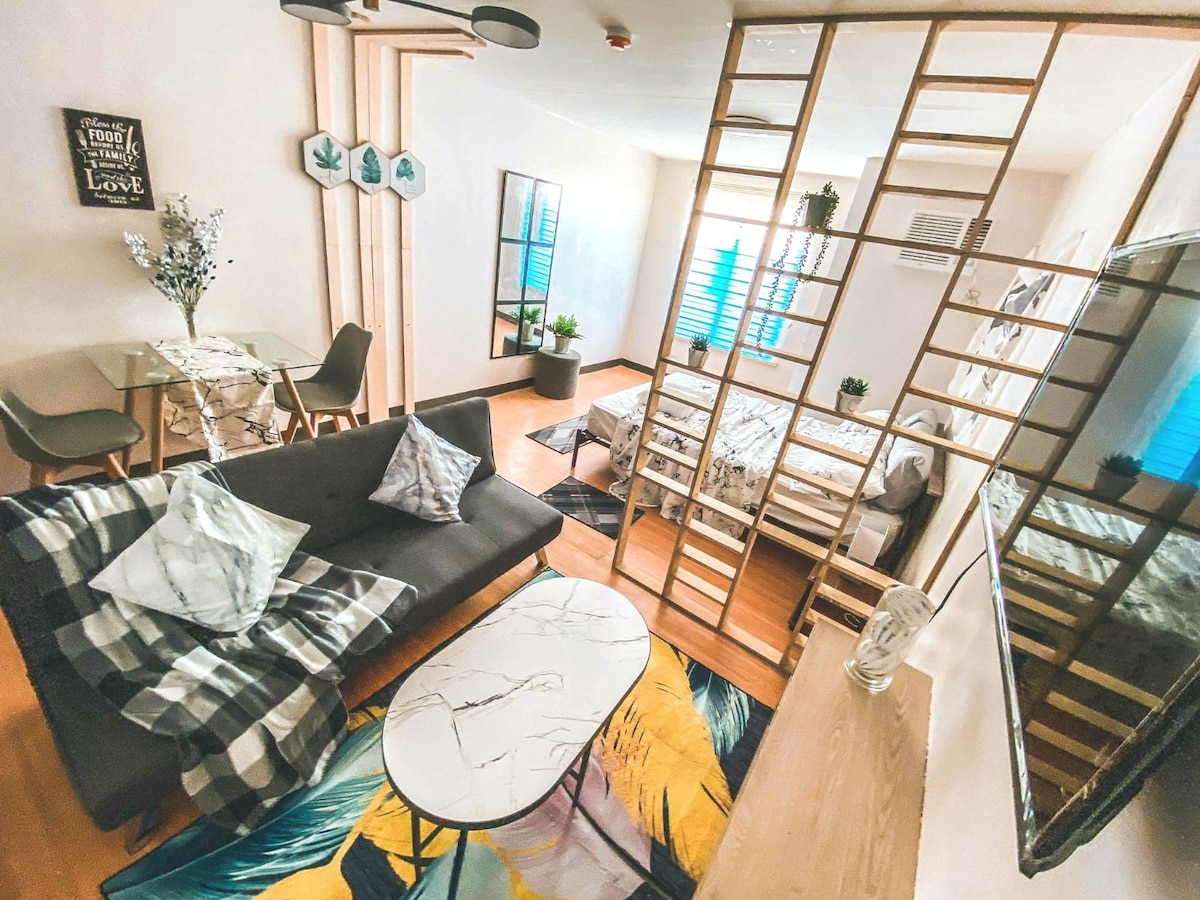 An open living area features a comfortable sofa with decorative pillows facing a stylish coffee table. A dining table for four is positioned beside a vibrant area rug. A partition separates the sleeping space, which is highlighted by natural light from blue curtains and surrounded by indoor plants.