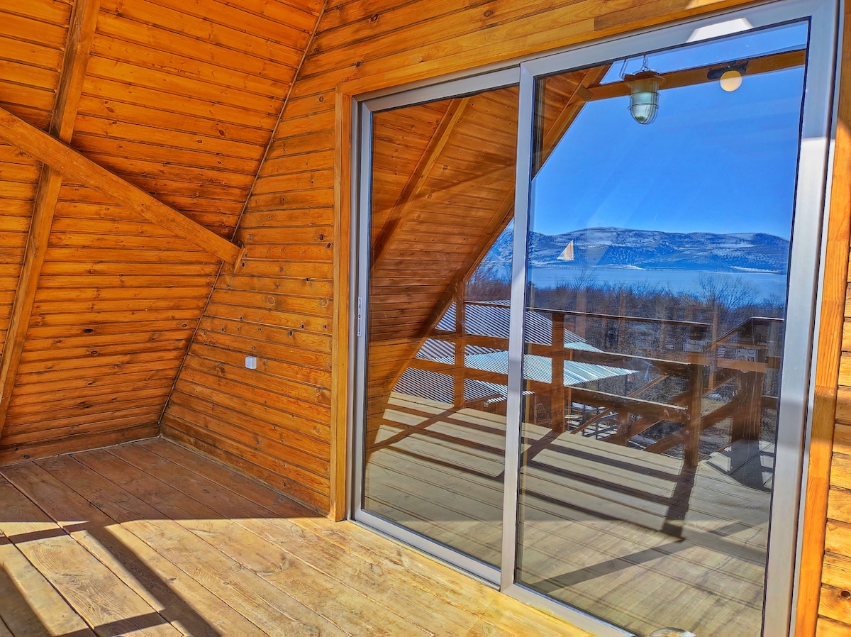 Large sliding glass doors reflect the surrounding landscape, contrasting against the warm wooden walls. The spacious balcony features wooden flooring, with a view of distant mountains and natural scenery visible through the glass panes.