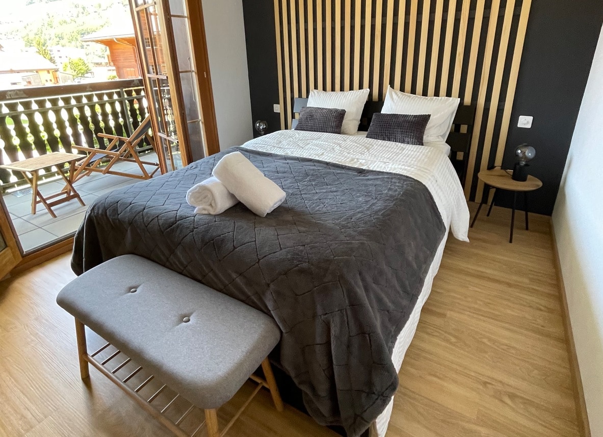 A cozy bedroom is featured with a spacious bed adorned with a textured gray blanket. A wooden accent wall adds character, while bedside tables provide additional lighting. A bench sits at the foot of the bed, and a large balcony door offers access to outdoor views.