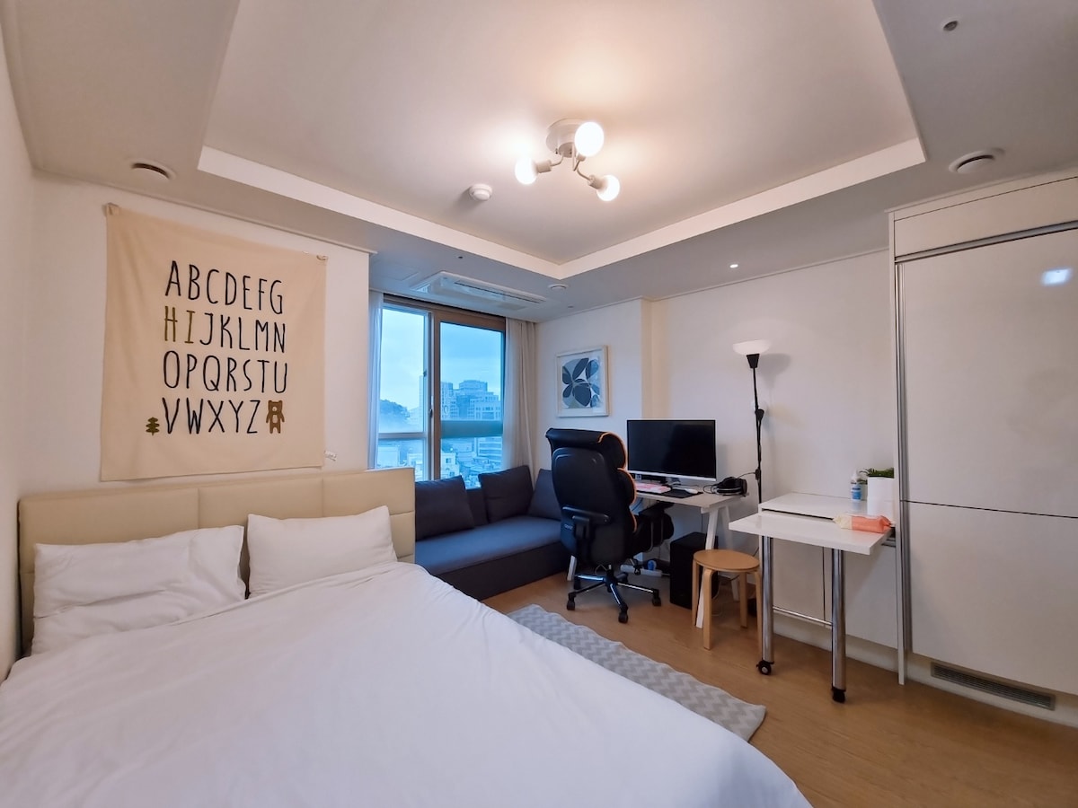 A well-organized room features a spacious bed with white linens and a wall adorned with alphabet art. A comfortable gaming chair and desk, equipped with a computer, are positioned near a large window offering city views. A small side table complements the cozy seating area.