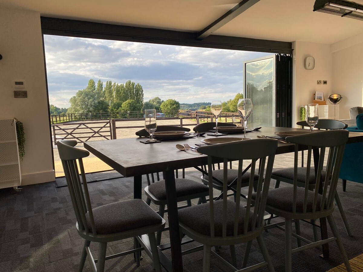 A dining table with dark wooden surface and six matching chairs is positioned in an open space. Large glass doors invite natural light and provide views of the rural landscape and courtyard. Nearby furnishings can be seen, adding to the welcoming ambiance.