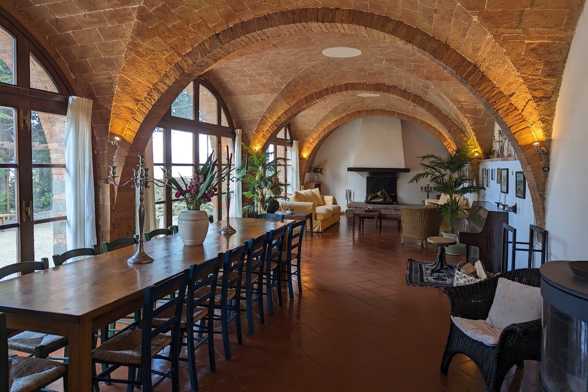 A spacious living and dining area features historic vaulted ceilings with warm lighting highlighting the stone arches. A large dining table accommodates multiple guests, while a cozy seating arrangement surrounds an open fireplace. Plants are positioned throughout, adding a touch of greenery to the space.