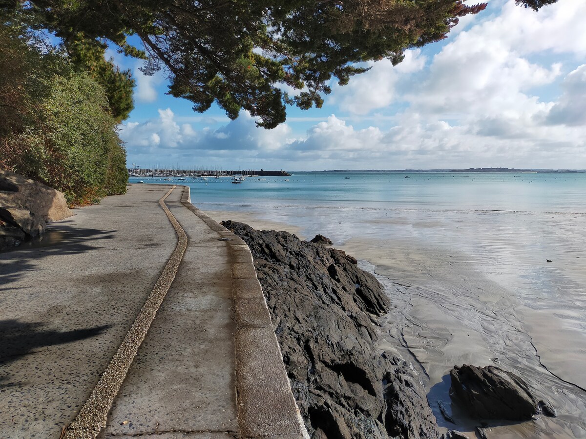 A coastal pathway runs alongside the calm water, framed by trees on one side. The water reflects a range of blue hues under a partly cloudy sky. Smooth rocks are visible along the shore, with boats gently bobbing in the distance.