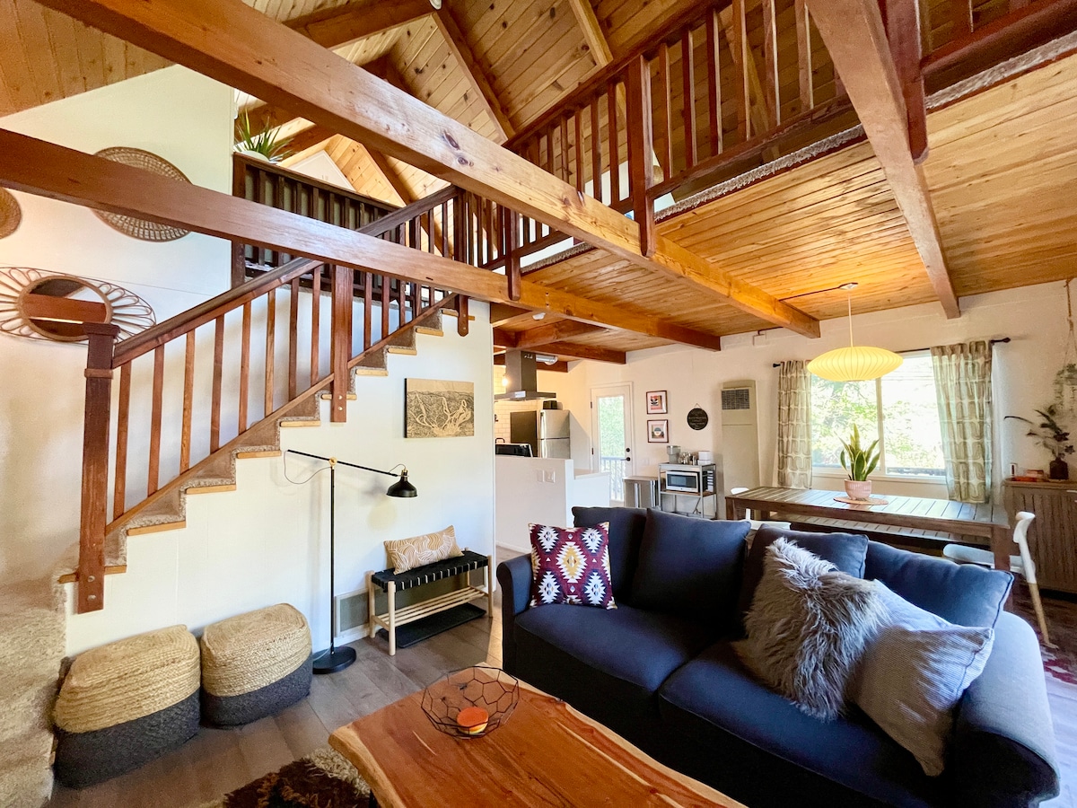 An open living area showcases rustic wooden beams and a staircase leading to a loft, which enhances the spaciousness. A dark sofa with decorative pillows is centered in the room, while natural light filters in through the windows, illuminating the cozy interior.