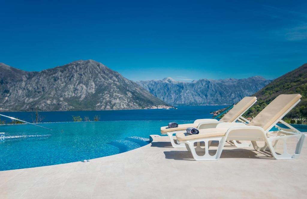 A serene infinity-edge pool is visible, blending with the surrounding landscape and offering expansive views of the Bay of Kotor and majestic mountains. Two lounge chairs are positioned at the pool's edge, inviting relaxation under clear blue skies.