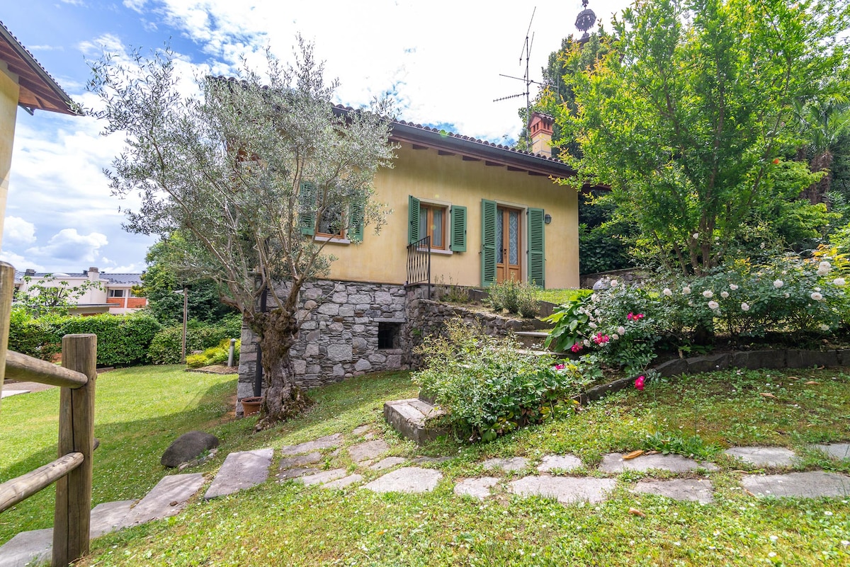 A charming villa is nestled within a vibrant garden, featuring a combination of greenery and blooming flowers. The stone pathway leads to the entrance, with a rustic facade and shuttered windows adding character. Soft sunlight illuminates the surroundings, enhancing the tranquil atmosphere.