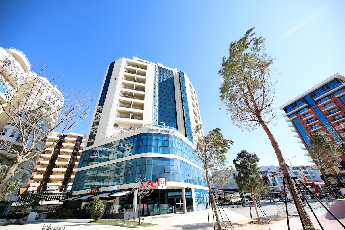 A modern multi-story building is prominently displayed, featuring large glass windows reflecting the clear blue sky. Surrounding greenery and nearby structures create a vibrant urban setting. The ground floor includes a recognizable fast food restaurant, contributing to the lively atmosphere.