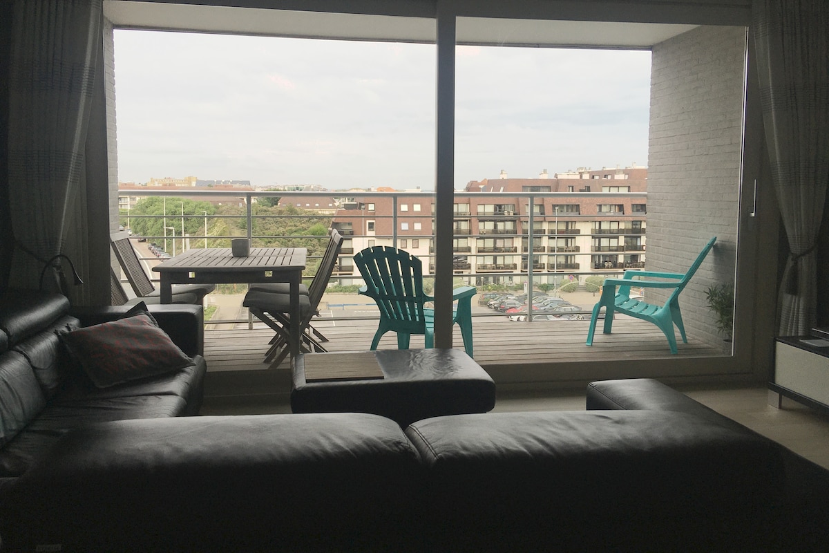 A spacious living area is presented with a black leather sectional sofa, looking out through large glass doors to a private terrace. An outdoor dining table and vibrant blue chairs are visible, along with views of the surrounding open area and nearby buildings.