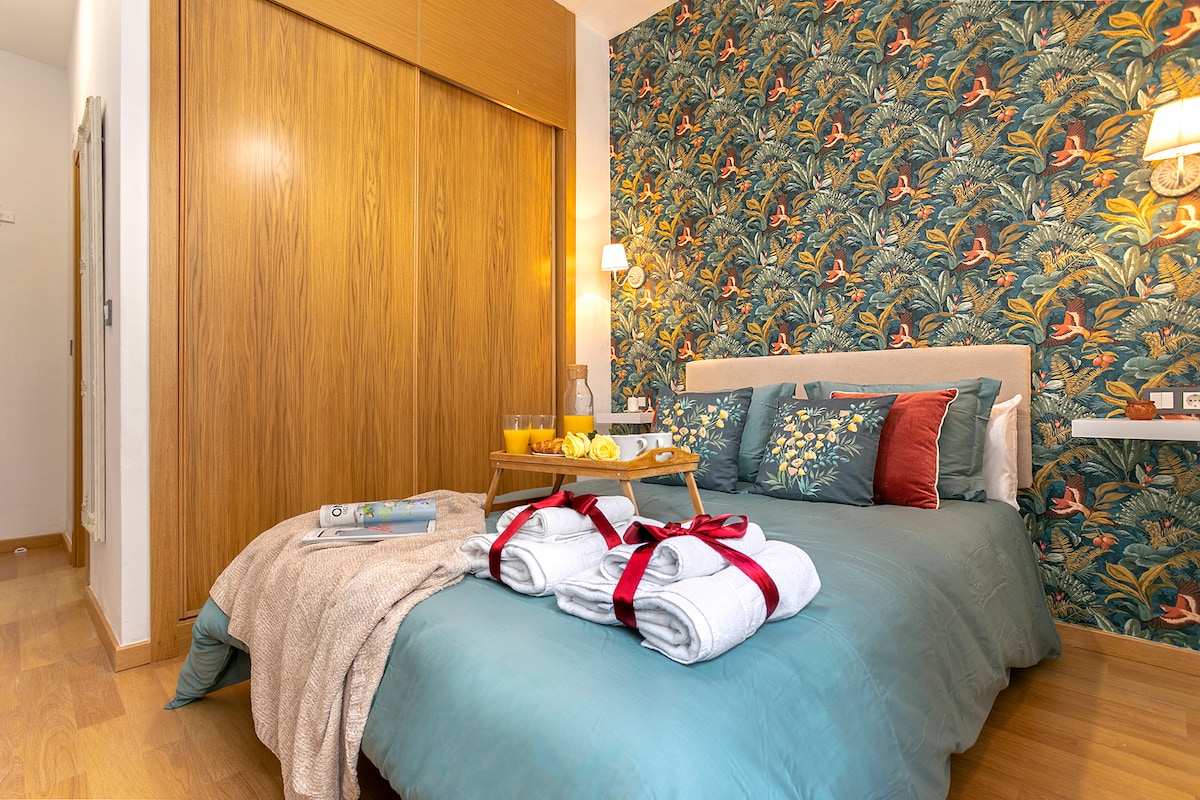 A bedroom is presented with a spacious bed adorned in teal bedding with decorative pillows. The wall behind features a vibrant floral pattern. A tray with fruit and beverages rests on the bed, while neatly arranged towels are placed at the foot, contributing to the room's welcoming ambiance.