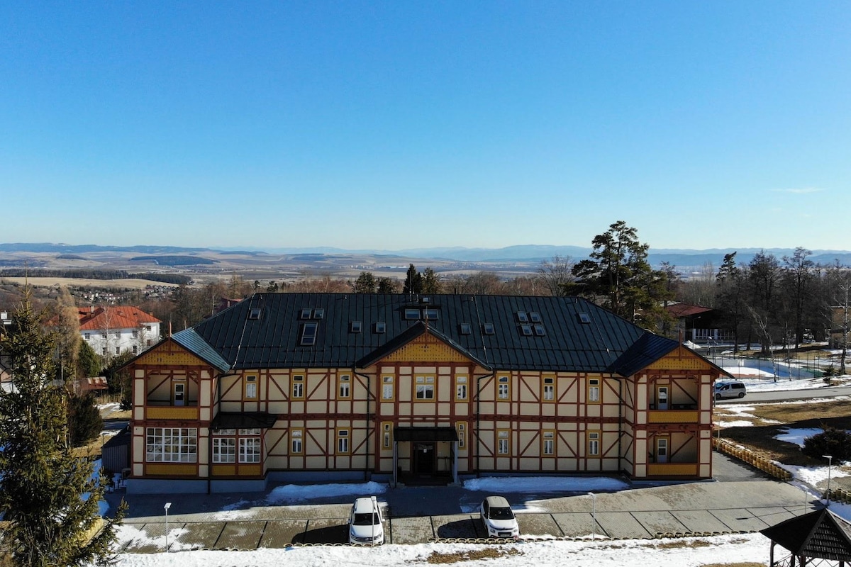The exterior of a historic building, featuring a charming wooden structure with a yellow and red facade. It is set against a clear blue sky, with snow partially covering the ground. Surrounding greenery and distant mountains can be seen in the background.