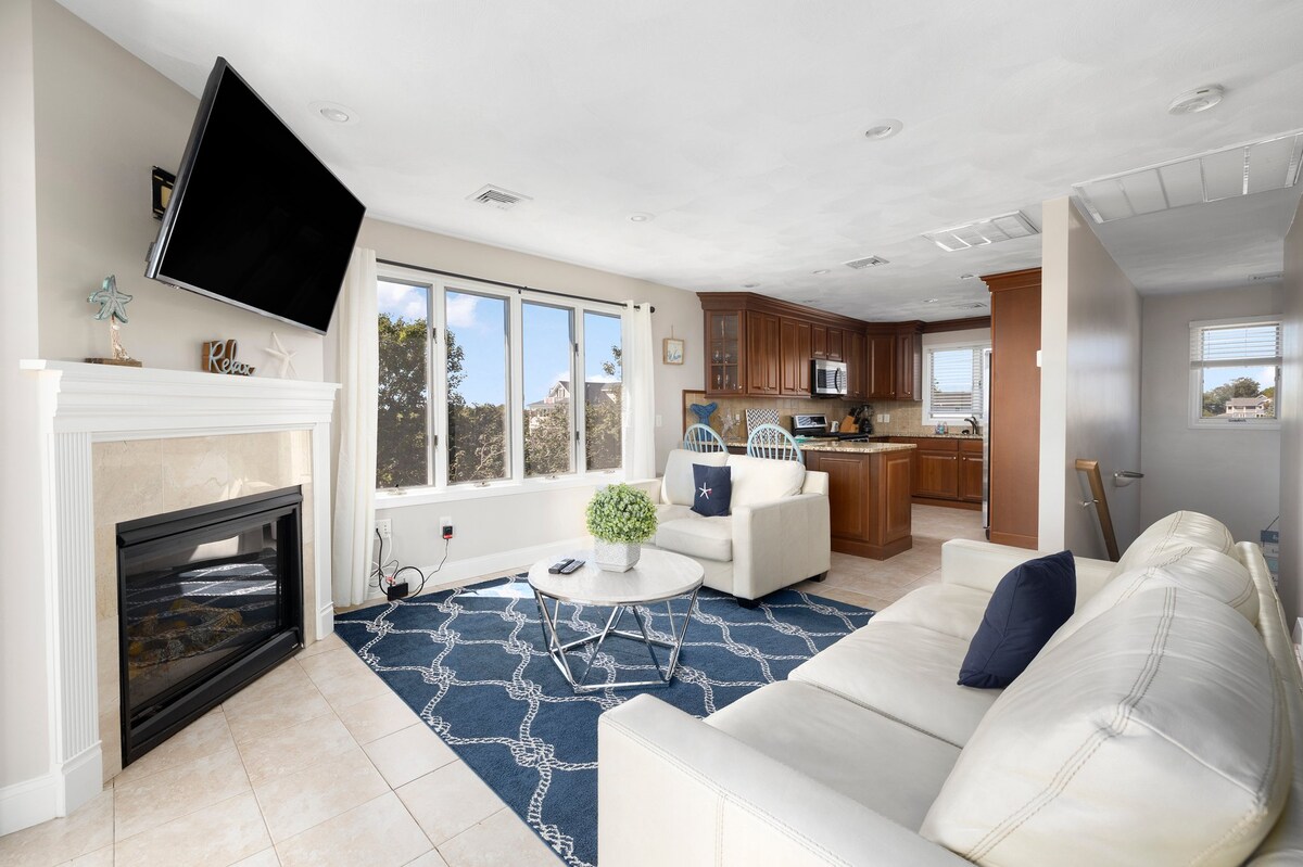 A bright living area is featured with a white leather sofa and accent chairs. A blue area rug adds a touch of color beneath the coffee table. Large windows allow natural light to fill the space, revealing a view of the exterior greenery. A modern kitchen is visible in the background.