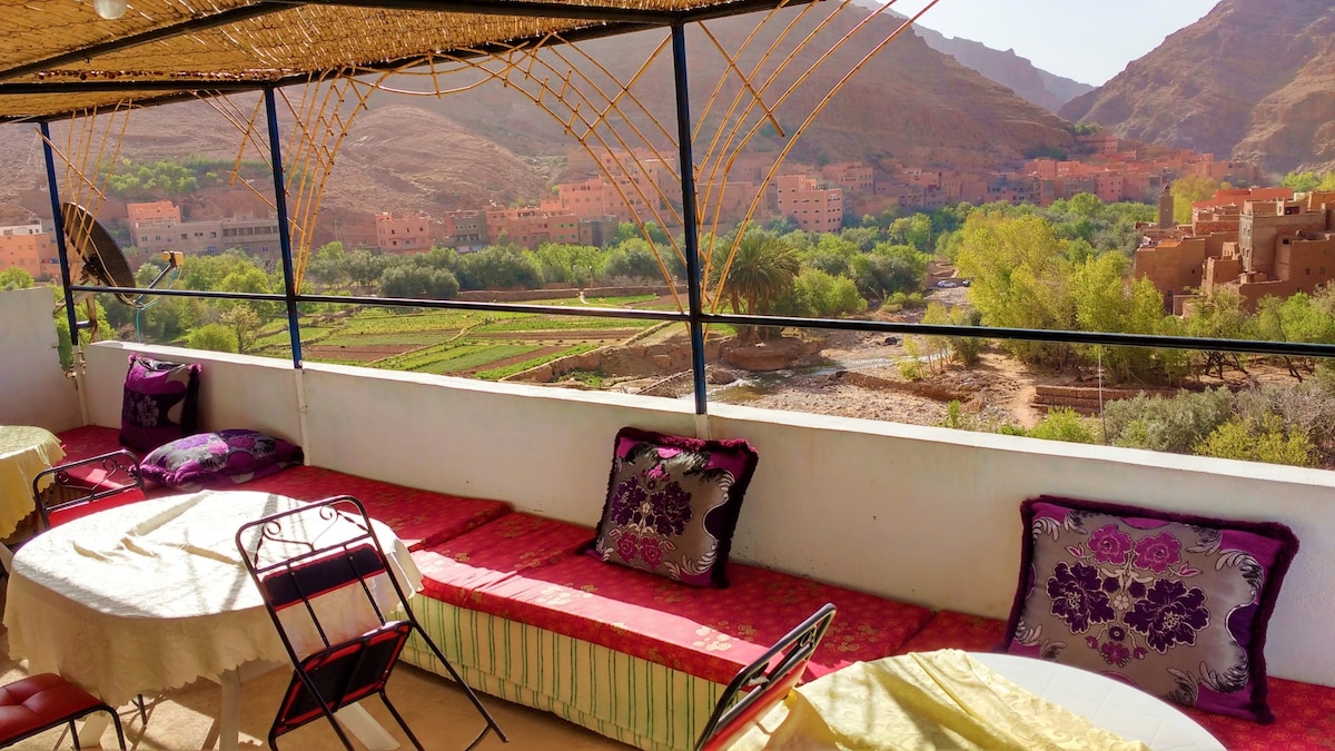 A spacious terrace features seating arrangements with colorful cushions and tables positioned for leisure. The backdrop includes mountains and greenery, creating a serene view over the landscape. Sunlight enhances the inviting atmosphere, while a canopy provides shade.
