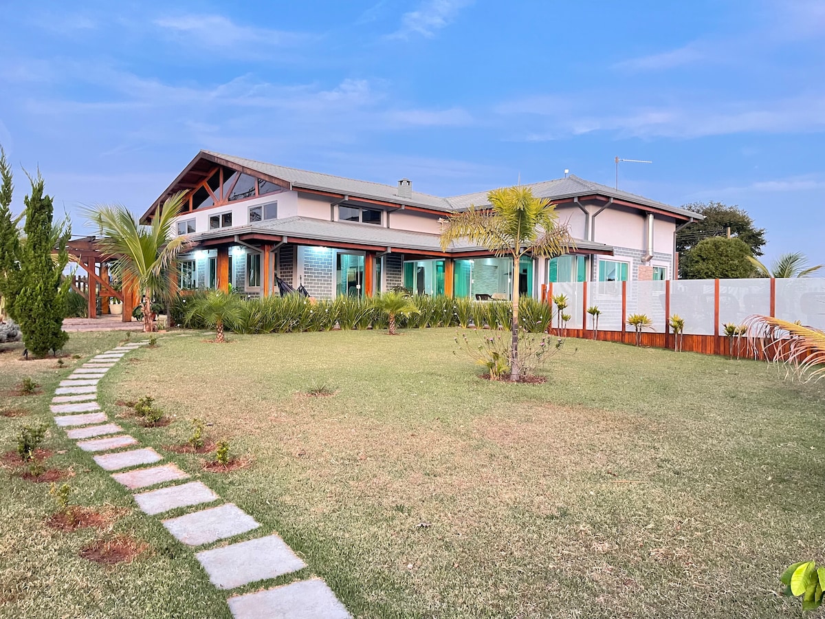 The exterior of a spacious two-story house is depicted, surrounded by a well-maintained garden. A stone pathway leads to the entrance, lined with lush greenery and palm trees. The house features large windows and a distinctive roof design, combining elements of modern architecture.
