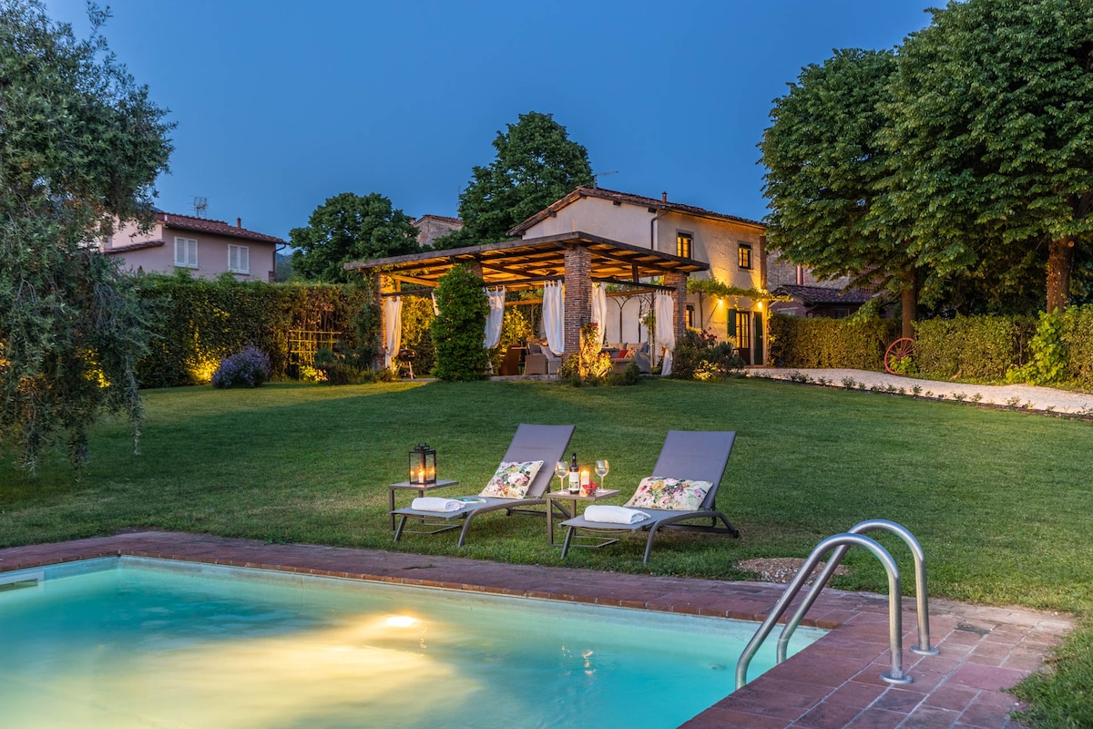 A private swimming pool is surrounded by a manicured lawn, featuring two lounge chairs with decorative pillows. A gazebo is visible in the background, offering a shaded area. Warm lights illuminate the garden and the villa, creating an inviting evening ambiance.