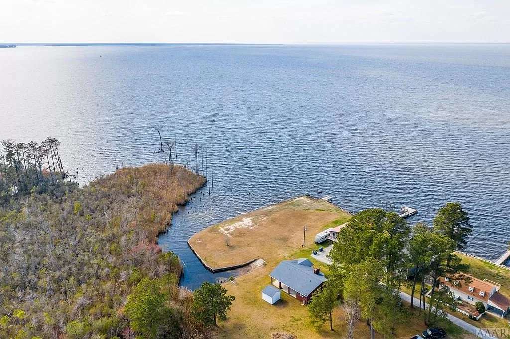 An aerial view captures a serene landscape with a vast body of water stretching to the horizon. A small grassy area is framed by trees, with a house positioned nearby. The calm waters are bordered by a wooded shoreline, emphasizing the tranquility of the location.