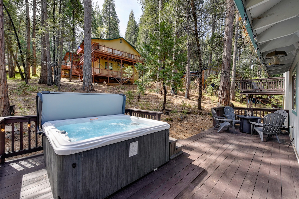 A spacious outdoor deck features a hot tub surrounded by forested scenery. Chairs are positioned nearby, creating a relaxing atmosphere for outdoor enjoyment. In the background, additional cabins are nestled among the trees, enhancing the natural setting.
