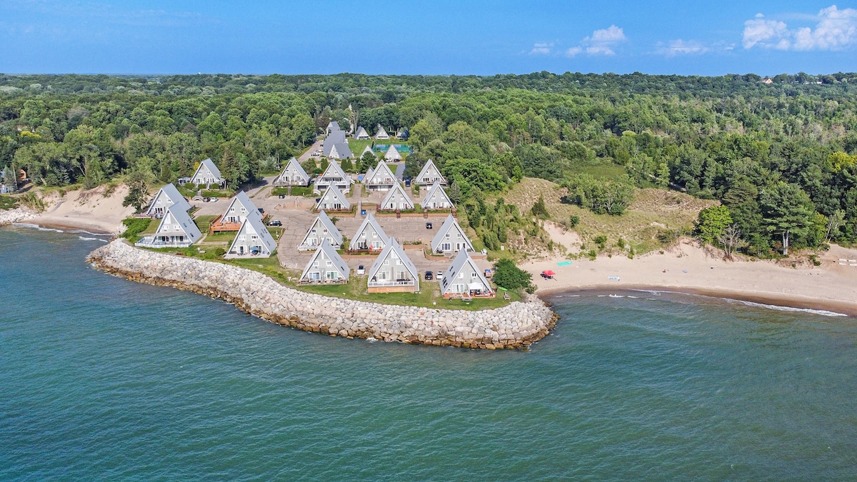 The image captures a serene aerial view of the chalet community situated along the shoreline of Lake Michigan. A series of triangular-roofed chalets are nestled among lush greenery, with sandy beach access visible in the foreground.