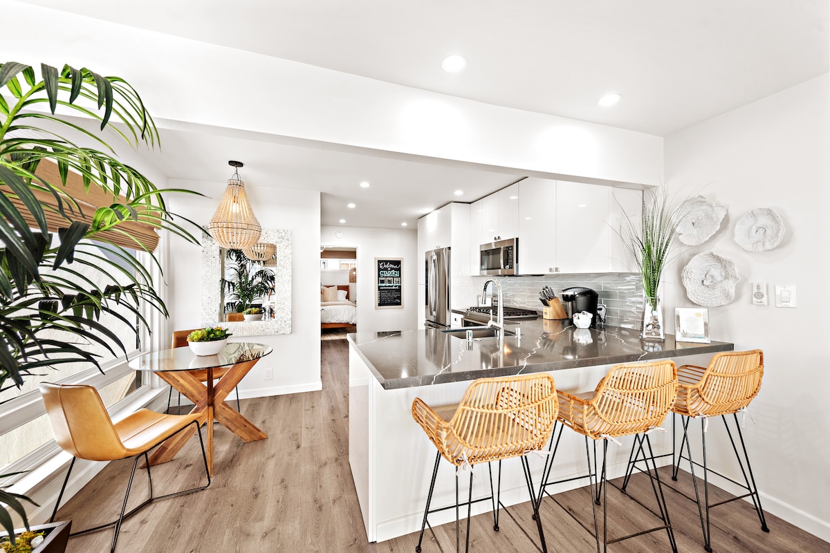An open kitchen and dining area features modern appliances and a spacious island with four woven bar stools. A round dining table is positioned nearby, complemented by potted greenery. Light-colored cabinets and countertops enhance the bright, airy atmosphere.