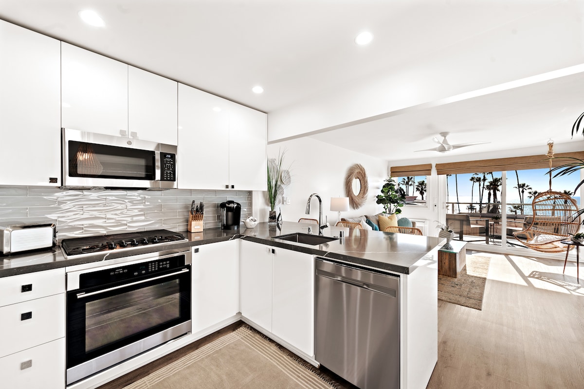 A modern kitchen is presented with white cabinetry and sleek countertops, featuring stainless steel appliances, including an oven, microwave, and dishwasher. A coffee maker and a decorative plant add to the inviting space. Natural light streams in from the open living area beyond.