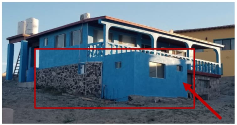 The exterior of a colorful building is visible, featuring a combination of smooth blue paint and stone accents. A spacious balcony with decorative railings extends along the upper level. Large windows provide natural light to the interior, while a staircase leads to the upper entrance.