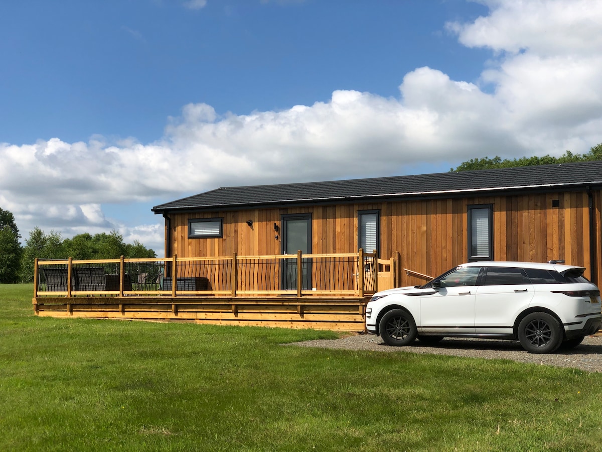 A spacious wooden lodge is set against a backdrop of grassy fields under a partly cloudy sky. A large sun deck extends from the lodge, with French doors visible, while a car is parked nearby on a gravel surface.