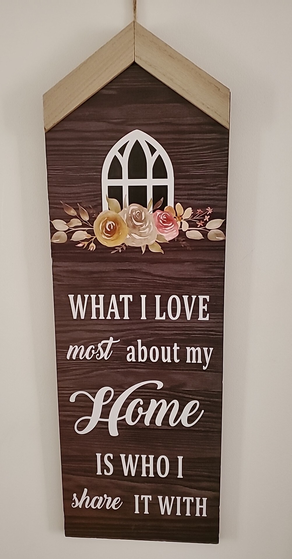 A decorative wall art piece features a wooden design and a roof-like top. Text in a mix of bold and cursive font states, 'What I love most about my home is who I share it with.' Decorative flowers are illustrated near the top, enhancing the overall design.