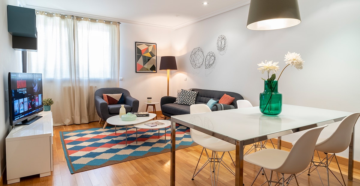 The living area features a modern design with a dark blue sofa and light-colored armchair. A round coffee table sits on a colorful geometric rug, complemented by a dining table with white chairs. Natural light filters through sheer curtains, enhancing the welcoming ambiance.