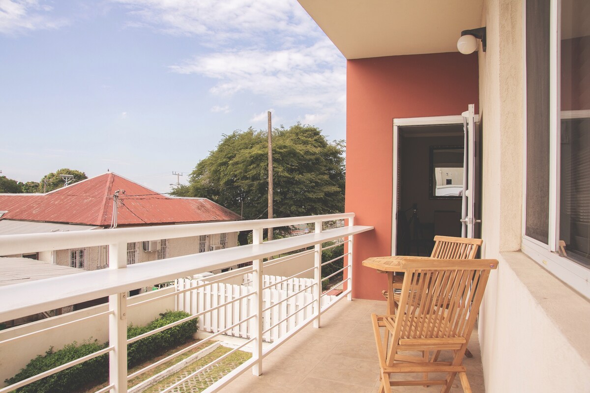 A private balcony is showcased with wooden seating positioned beside a view of adjacent greenery and neighboring rooftops. The relaxing outdoor space is bordered by a sleek railing, adding to the contemporary feel of the apartment.