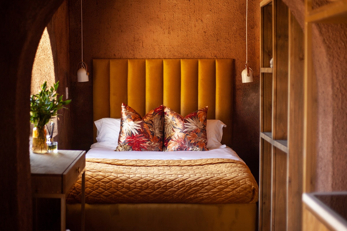 A cozy sleeping area is highlighted by a yellow velvet headboard and decorative pillows featuring a tropical pattern. Soft bedding layers are complemented by a warm-toned coverlet. Natural light enters through an archway, enhancing the earthy texture of the surrounding walls.