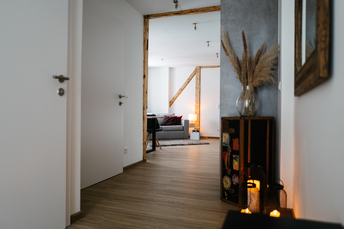 A well-lit hallway leads to a living area with a comfortable sofa. Decorative elements, such as plants and a lantern, are placed along the walls, contributing to the home's cozy ambiance. Warm wooden beams are visible, enhancing the stylish interior.
