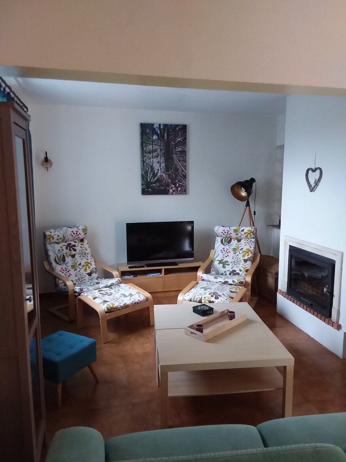 A cozy living area is arranged with two patterned armchairs and a small teal stool. A wooden coffee table sits in the center, complemented by a flat-screen television mounted on the wall. Soft light spills in from a nearby lamp, enhancing the inviting ambiance.