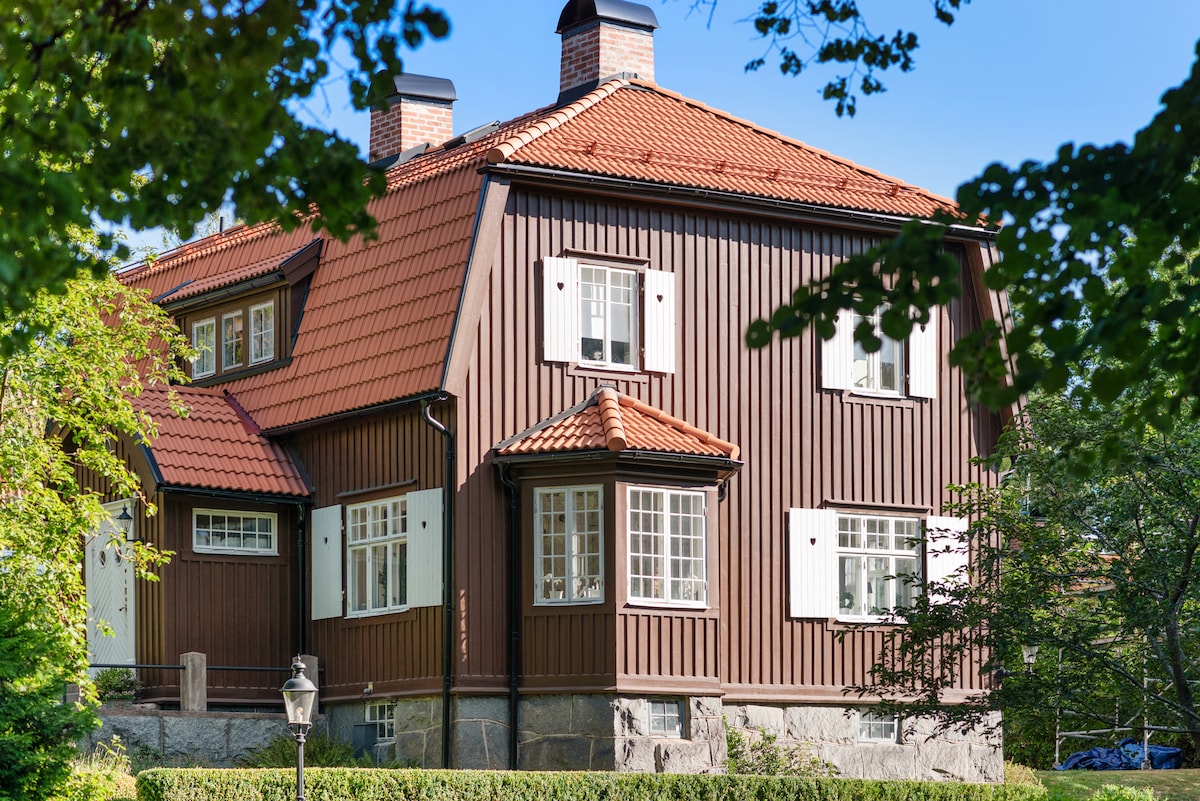 A two-story wooden house features a warm brown exterior with a red-tiled roof. Bright white window frames and shutters add contrast, while landscaped greenery surrounds the property. A stone pathway leads to the entrance, enhancing the inviting atmosphere of this historic home.