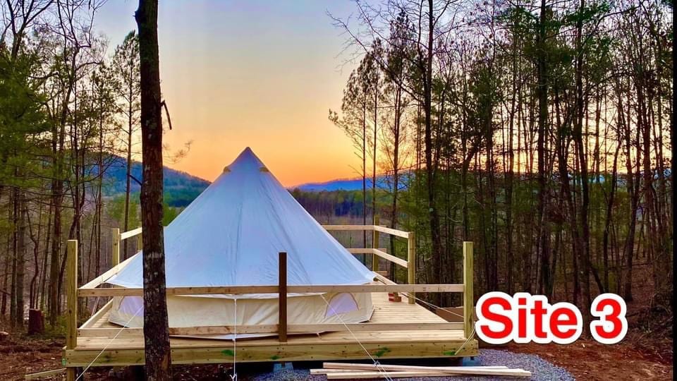 A glamping tent is situated on an expansive wooden deck, surrounded by trees with a scenic view of a sunset in the background. A pathway leads up to the tent, emphasizing the serene, natural setting at Site 3.