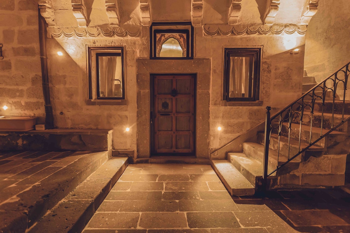 Warmly lit stone steps lead to a wooden door, framed by intricately carved archways. Flanking the entrance are windows draped in light fabric, enhancing the inviting ambiance of the entrance area. Soft lighting reflects off the textured stone walls, highlighting the architectural details.
