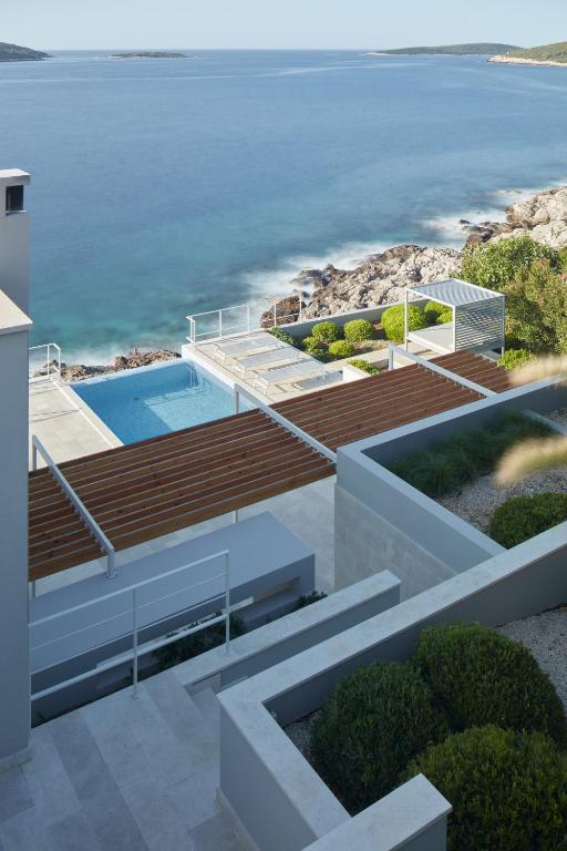 A view of the tranquil sea and rocky coastline is framed by a well-maintained garden. A swimming pool is situated beside a wooden deck with sun loungers, offering an inviting space for relaxation. The modern villa design complements the serene coastal landscape.