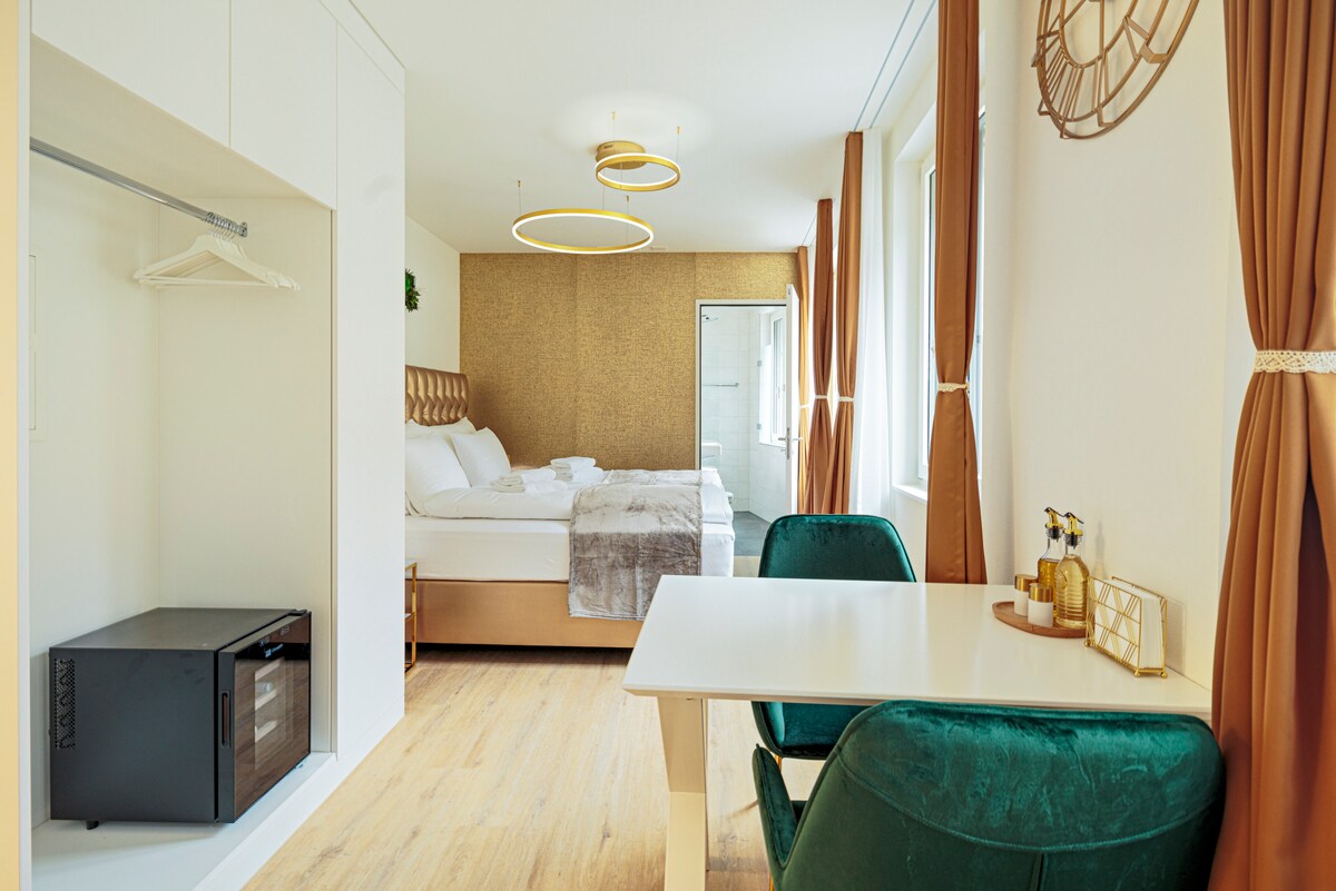 A spacious superior studio features three large windows allowing ample natural light. A comfortable kingsize bed is paired with a minimalist table and modern green chairs. A microwave is placed beside the closet, and a stylish decorative light fixture hangs from the ceiling.