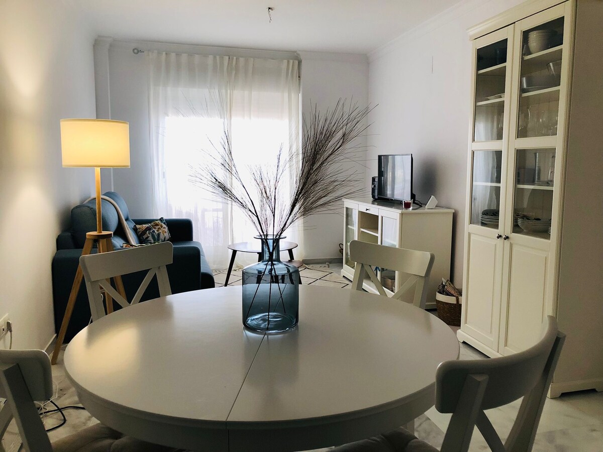 A light and airy living area is highlighted by a circular dining table and a glass vase with dried plant decor. A comfortable sofa is positioned near a window, providing natural light. A television is placed on a cabinet against the wall, completing the welcoming atmosphere.