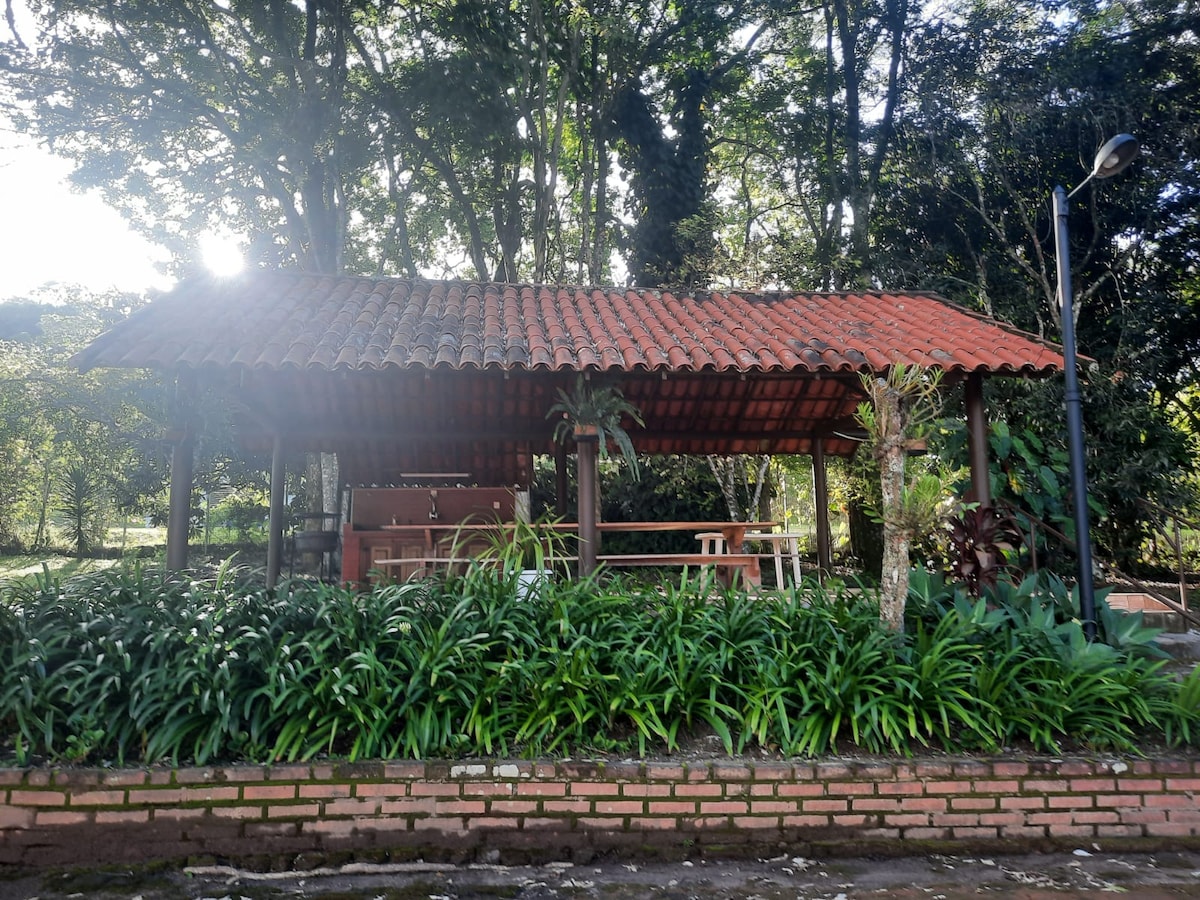 A rustic gazebo is surrounded by lush greenery, featuring a tiled roof and wooden tables. The open structure offers a barbecue area, illuminated by natural light filtering through the surrounding trees.