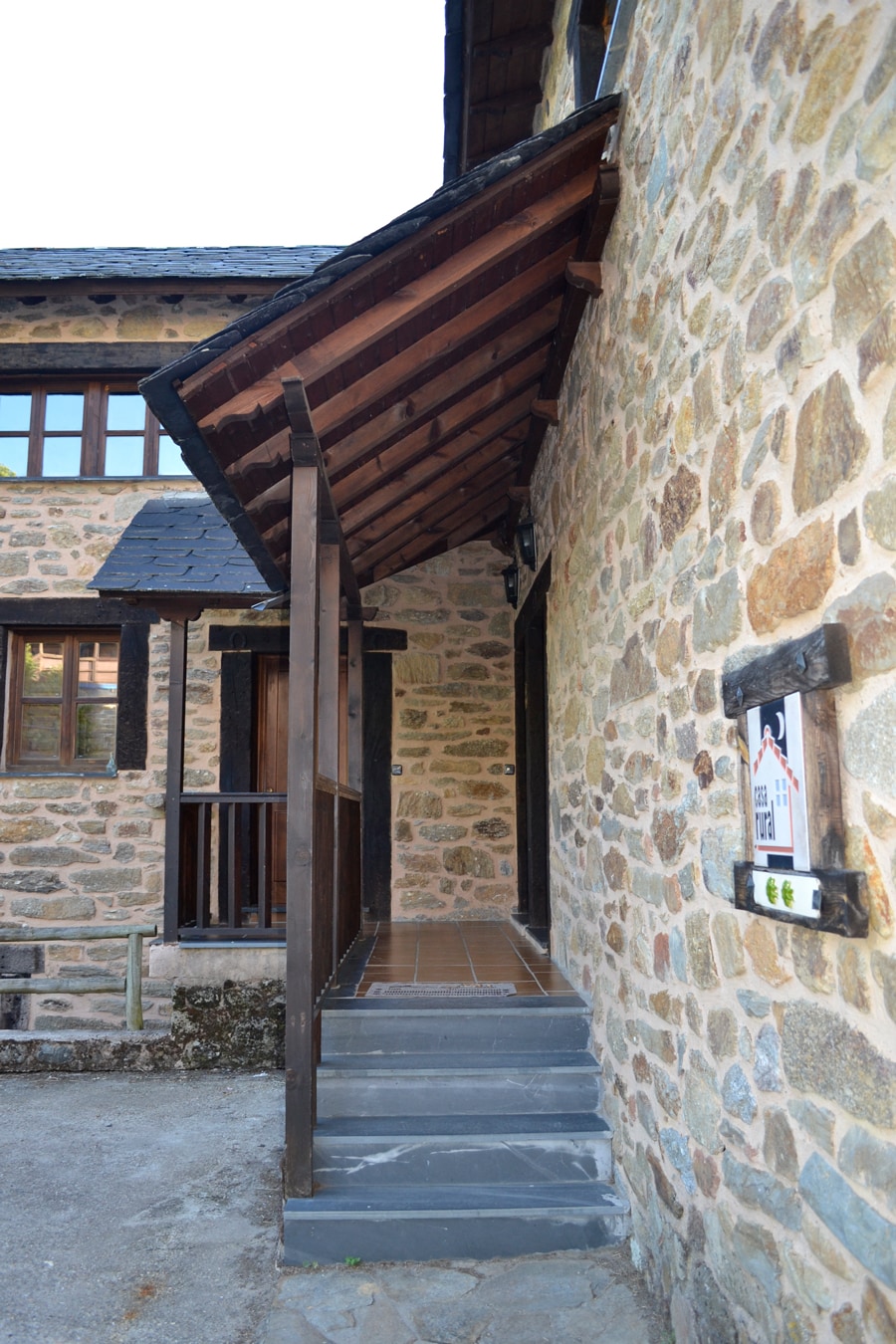 An entryway is framed by a wooden overhang, leading to a stone facade. Steps ascend towards the entrance, flanked by wooden beams. The rustic charm of the structure is complemented by natural elements in the surroundings.