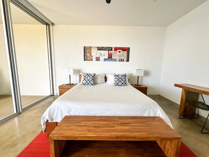 - Acogedora cama doble con muebles de madera sencillos pero elegantes y al lado de lámparas que crean un ambiente cálido.
- Las hermosas obras de arte agregan un toque de color y personalidad a la habitación.
- Las grandes puertas de vidrio ofrecen abundante luz natural y un fácil acceso al espacio al aire libre.